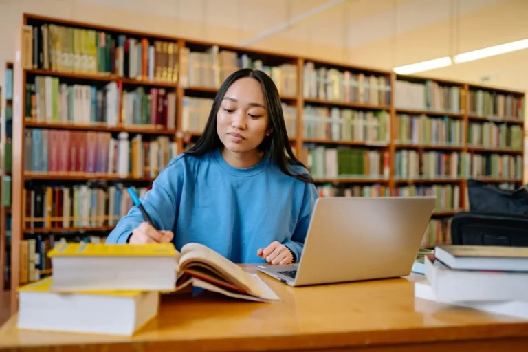 Joven estudiando en una biblioteca con libros y portátil