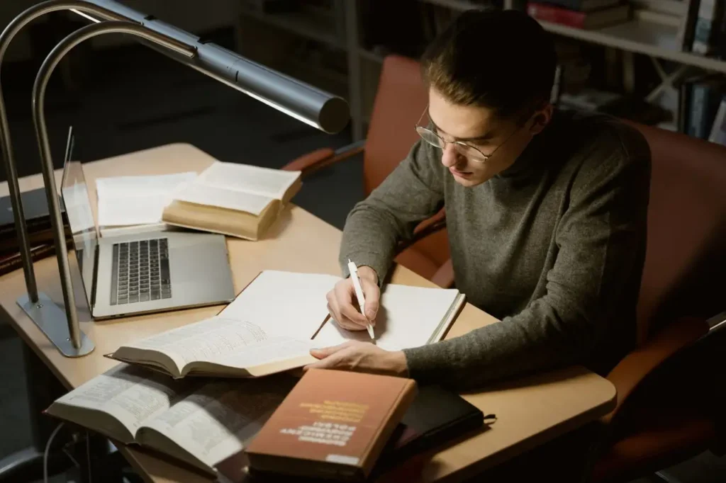 Estudiante tomando apuntes con libros y portátil bajo una lámpara
